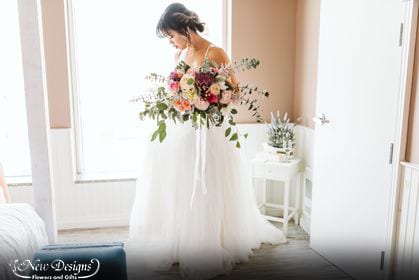 A Bride stands in front of a window, with soft lighting, looking down at a bouquet of Vibrant hues, White Peonies, Lavender fuji mums, Bright pink miniature roses, peach berries, blush ranunculus, with explosive stems of baby and silver dollar eucalyptus